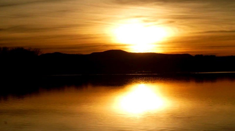 A golden sunset over distant hills is reflected on the calm surface of Lake LBJ, with dark silhouettes of the landscape contrasting against the bright sky and water near Lodge on the Lake.