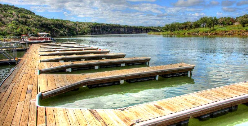 Wooden boat docks at Thunderbird Resort extend into a calm, clear lake, framed by green hills and trees beneath a blue sky with scattered clouds. The empty docks and tranquil water reflect the stunning landscape surrounding Thunderbird Resort.