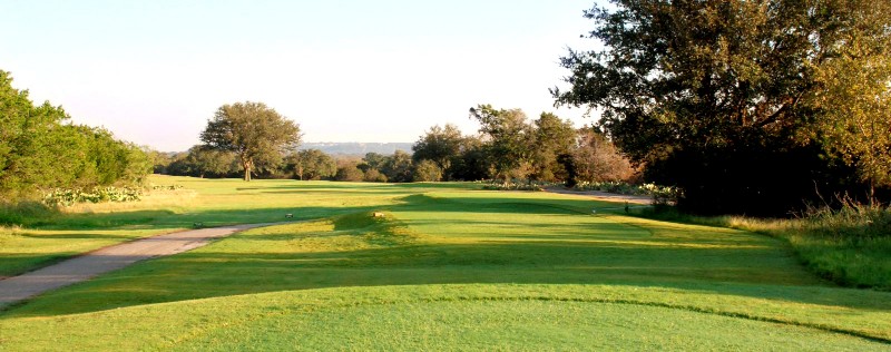 A green municipal golf course fairway at Delaware Springs, surrounded by trees under a clear sky, with a paved cart path running alongside the grass.