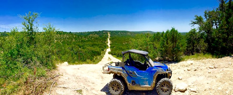 A blue off-road vehicle is parked on a rocky trail at Hidden Falls, overlooking a vast, green landscape with trees and hills beneath a clear blue sky.