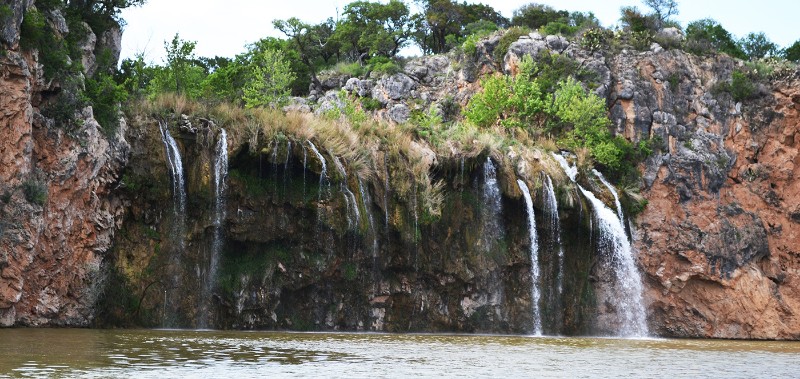 A rocky cliff at Lake Buchanan with multiple streams of water cascading into a murky pool below, surrounded by green shrubs and trees atop the cliff.