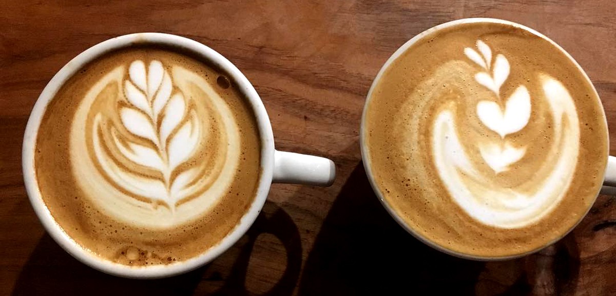 Two cups of specialty coffee with latte art rest on a wooden surface. The left cup shows a leaf design, while the right displays a similar, slightly asymmetrical leaf—both showcasing the craft of skilled coffee roasters.