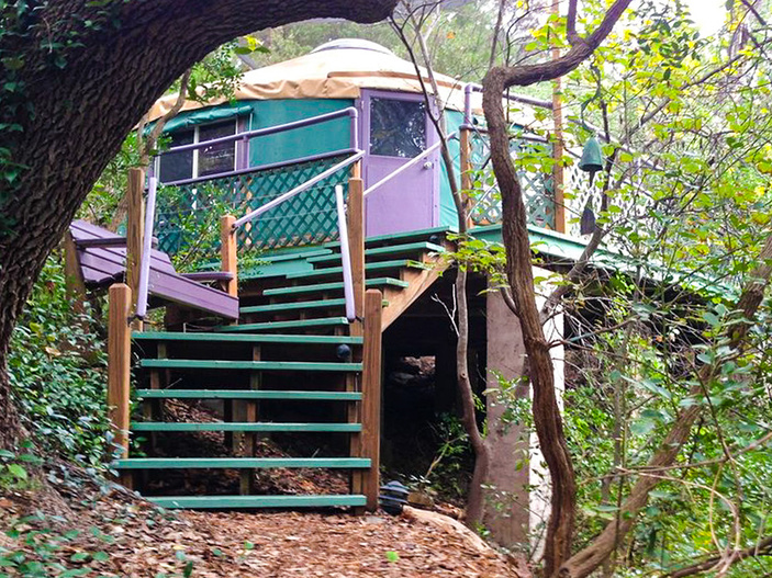 A green and purple yurt sits elevated on wooden stilts in a forest near Lake Buchanan, accessible by a staircase surrounded by trees and foliage at the charming Rainbow Hearth B & B.