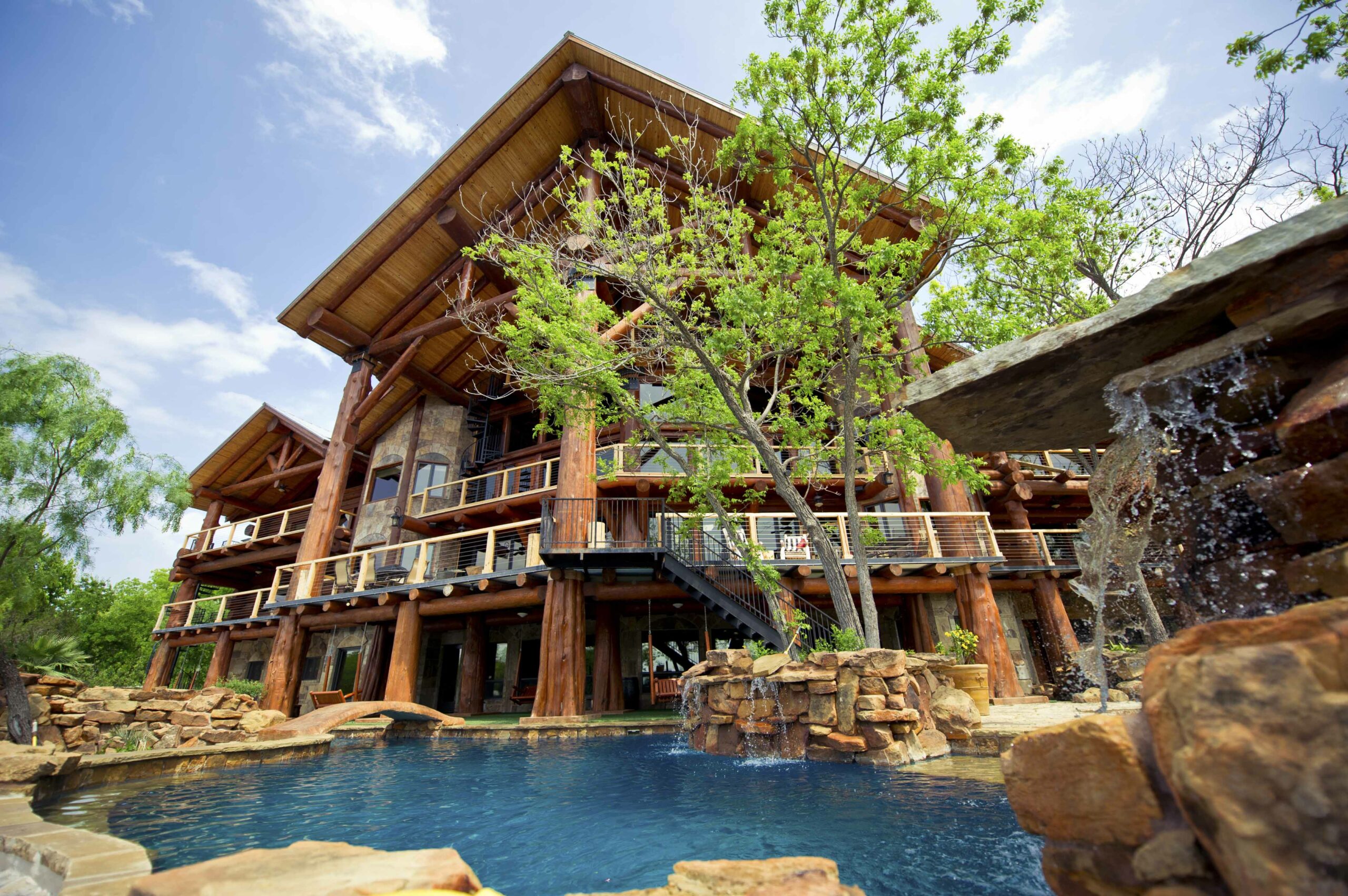 A large, rustic log cabin at Log Country Cove features balconies overlooking a blue pool with rocky edges and a small waterfall, surrounded by trees and greenery under a partly cloudy sky near Lake LBJ.
