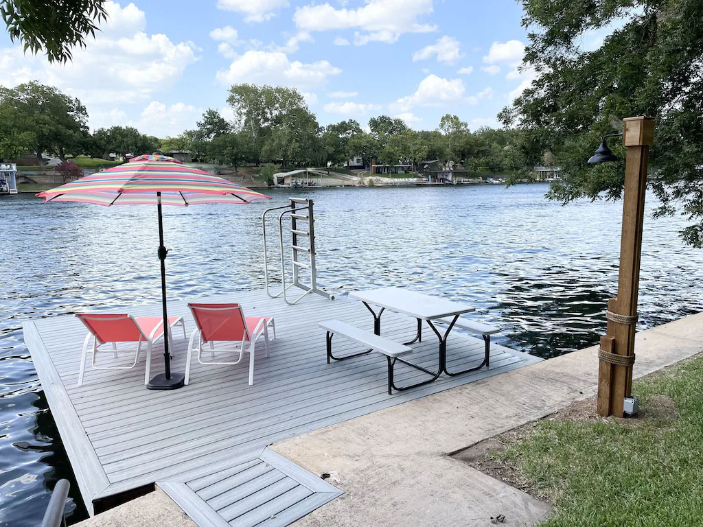 At Camp Clen Oak, a dock with two red lounge chairs under a striped umbrella and a white picnic table overlooks a calm river, surrounded by trees and houses under a partly cloudy sky.