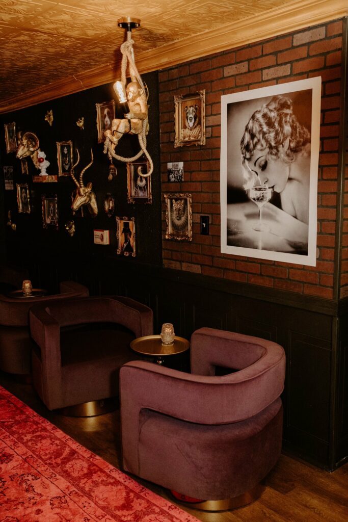 A cozy lounge at Sips On Second features two purple velvet chairs, a small round table, and eclectic wall art, including gold-framed portraits, monkey sculptures, and a large black-and-white photo of a woman sipping from a glass.