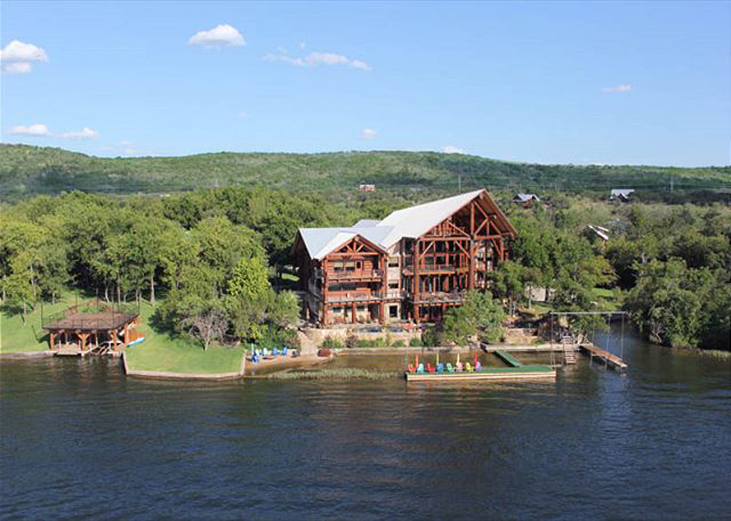 A large wooden lodge with a metal roof sits by the lake at Log Country Cove, surrounded by trees. Colorful kayaks and canoes line the shore near a dock that extends into the water, offering lakeside luxury beneath green hills and a blue sky.