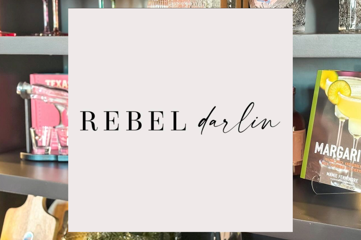 A light pink square with the words Rebel Darlin in bold and script fonts, set against shelves holding colorful books, glassware, and kitchen items. Rebel Darlin stands out stylishly amid the lively background.