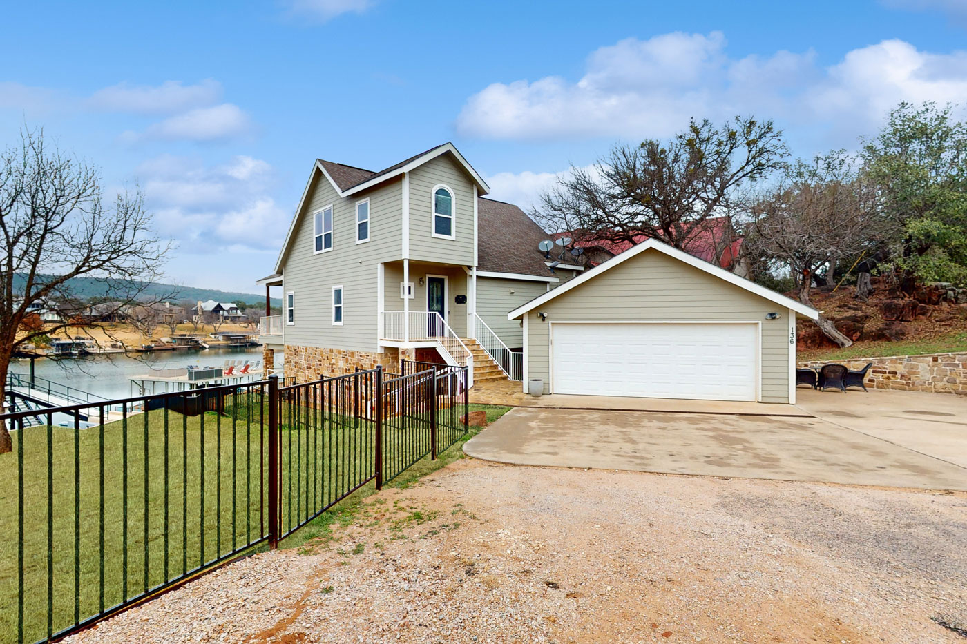 Two-story beige house with white trim and a stone foundation, featuring a double garage, front steps, and a fenced grassy yard overlooking the Still Waters lake, with trees and a clear sky in the background.