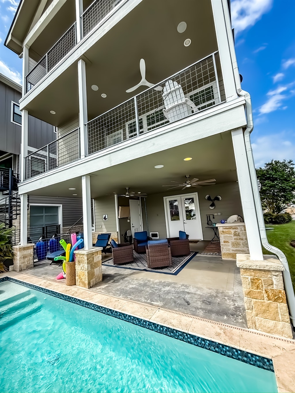 A modern three-story house with balconies overlooks a patio with outdoor seating and a swimming pool. Pool toys and towels are visible, and ceiling fans are installed on each balcony. The sky is partly cloudy.