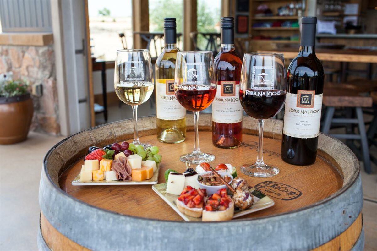 A wine barrel holds three glasses of wine, four bottles of assorted wine, and two plates with assorted cheeses, meats, fruit, and bruschetta, set in a rustic winery tasting area.
