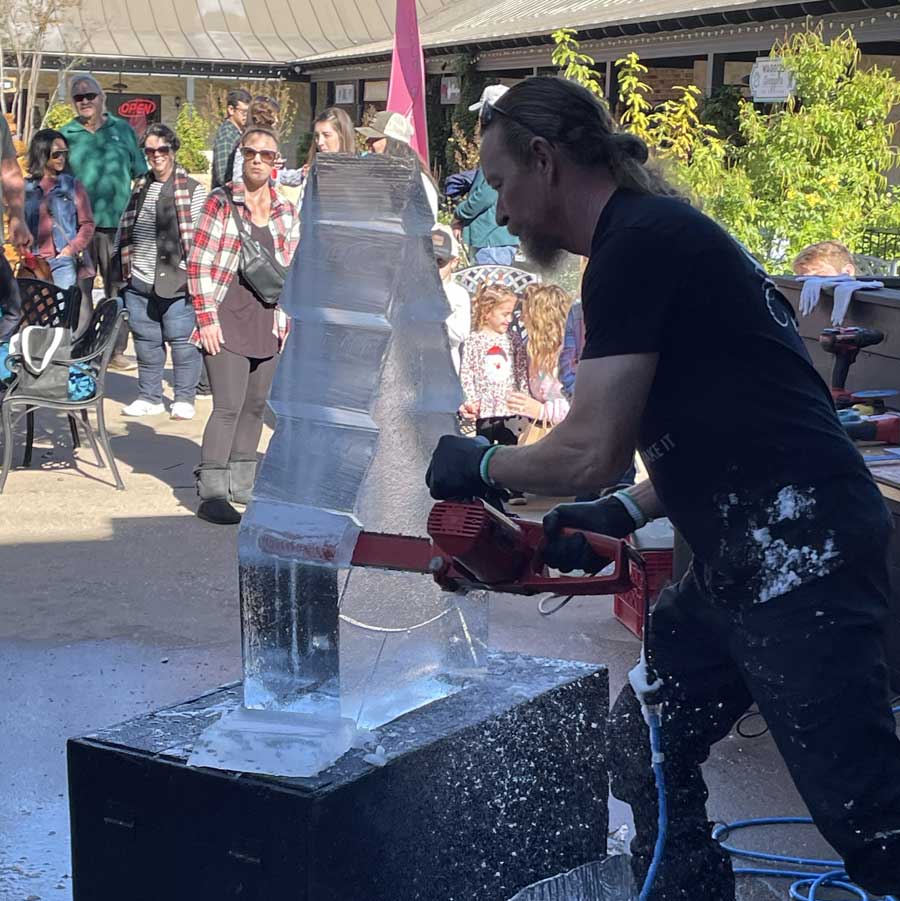 A man uses a chainsaw to carve an ice sculpture outdoors at the Highland Lakes Christmas Trail, while a crowd of people, including children, watch in the background on a sunny day.