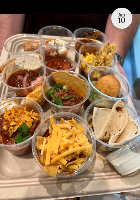 A tray holds nine small clear cups filled with various foods, including chili topped with cheese, crackers, chips, tortillas, and a fried ball, each cup with different garnishes and sides. A hand is visible underneath the tray.