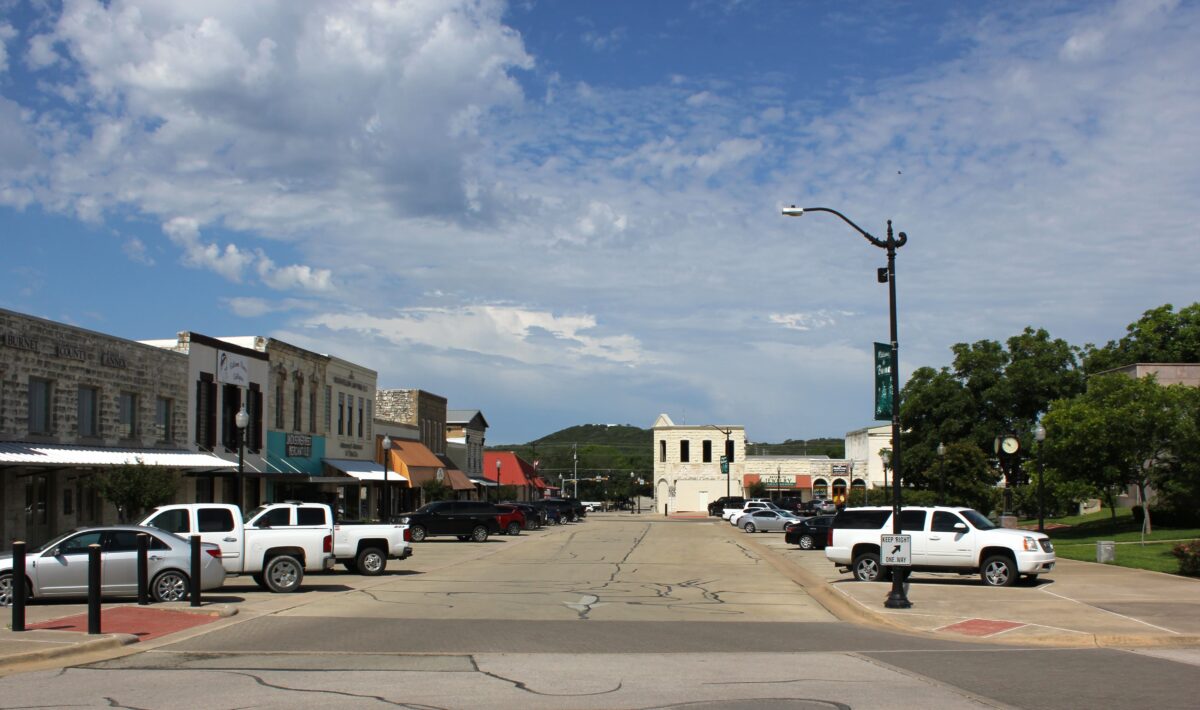 A small town main street with parked white pickup trucks, old storefronts with colored awnings on both sides, and a partly cloudy sky overhead. Trees and distant hills are visible in the background.