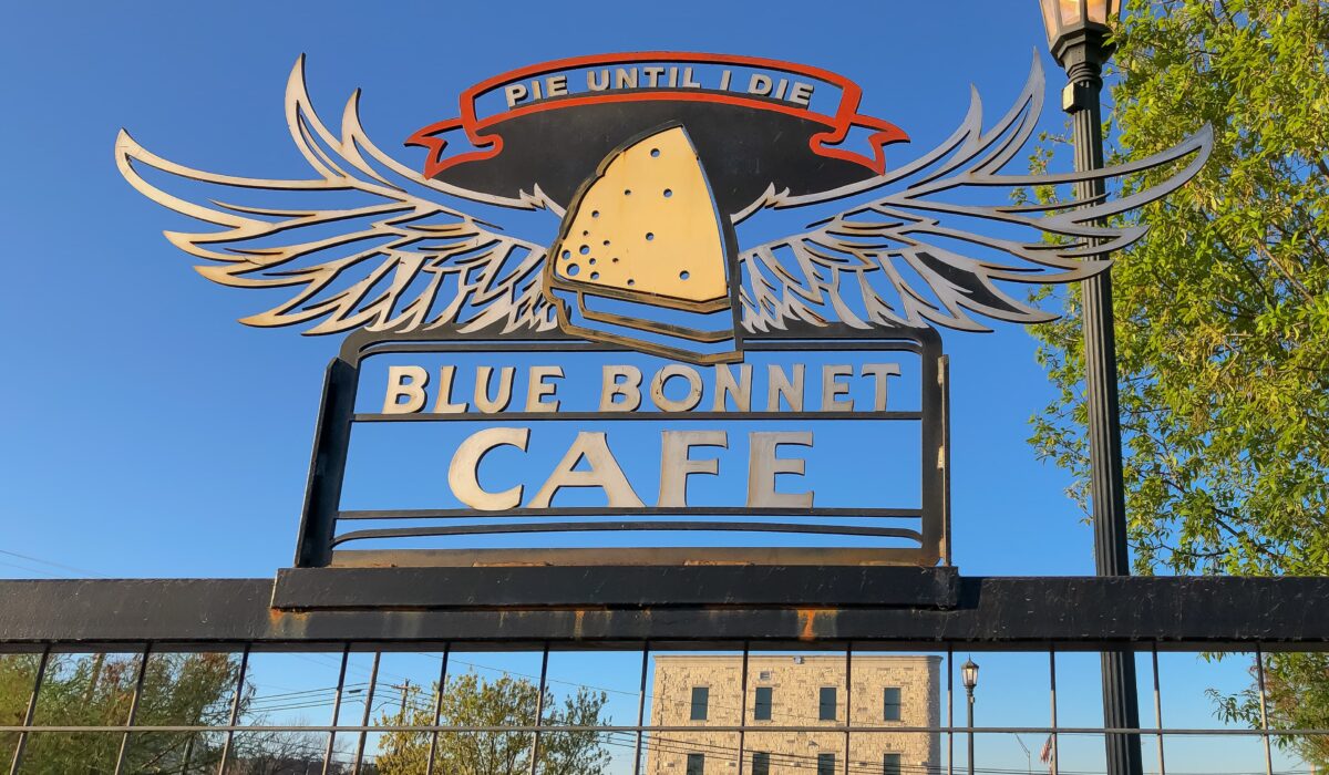 Metal sign for Blue Bonnet Cafe featuring a slice of pie with wings, a banner reading Pie Until I Die, and a clear blue sky in the background.