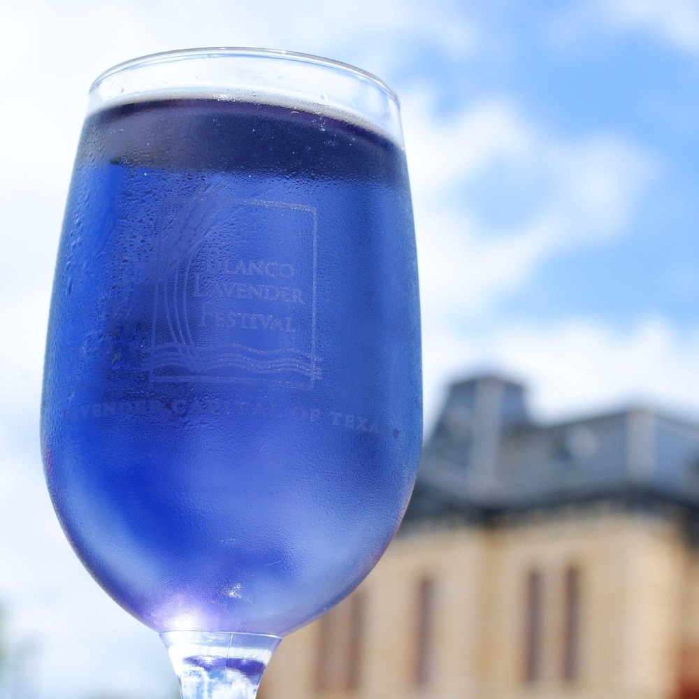 A wine glass filled with a vibrant purple drink, featuring the words Blanco Lavender Festival etched on it, held up against a bright blue sky and a blurred historic building in the background.