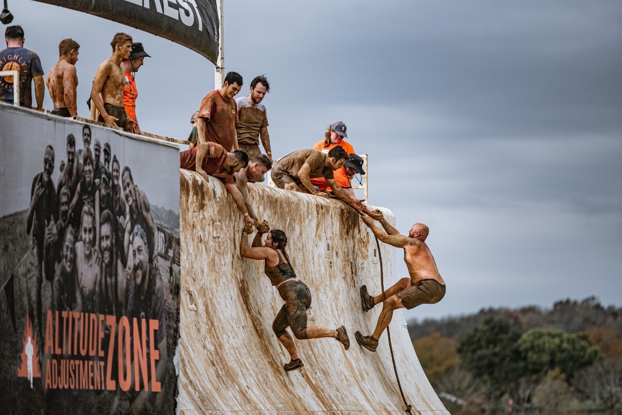 A group of muddy participants help each other climb a steep, slippery wall at an outdoor obstacle course event, with cloudy skies and trees in the background. A sign reads “Altitude Adjustment Zone.”.