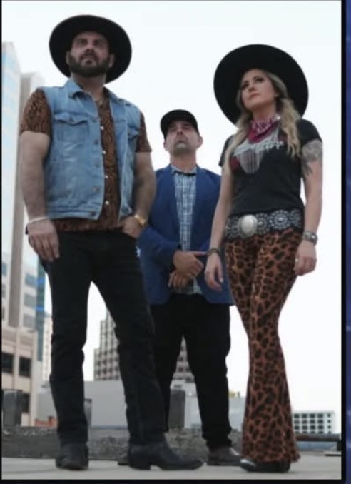 Three people wearing cowboy hats stand on a rooftop in a city. One wears a denim vest and leopard shirt, another in a blue jacket, and a third in a bandana, black top, and leopard print pants. Buildings are visible in the background.