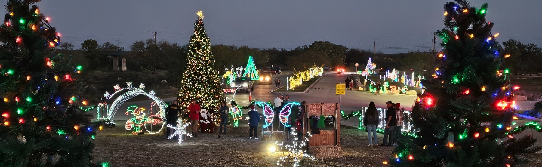A festive outdoor holiday display at dusk features a large decorated Christmas tree, illuminated arches, and various light sculptures along a road, with people admiring the decorations and trees wrapped in colorful lights.