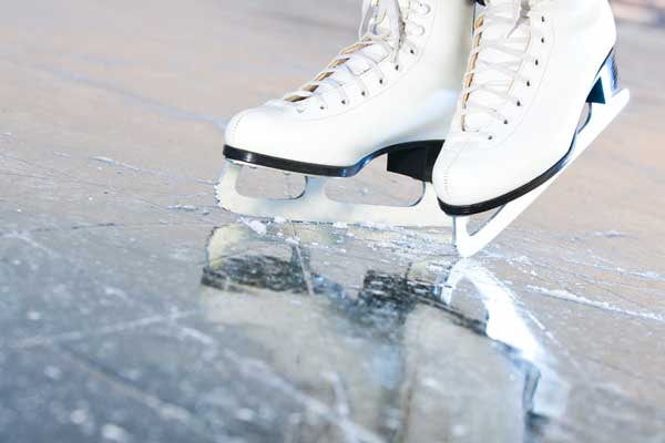 Close-up of a pair of white figure skates gliding on an ice rink, with the blades creating scratches and a reflection on the smooth ice surface.
