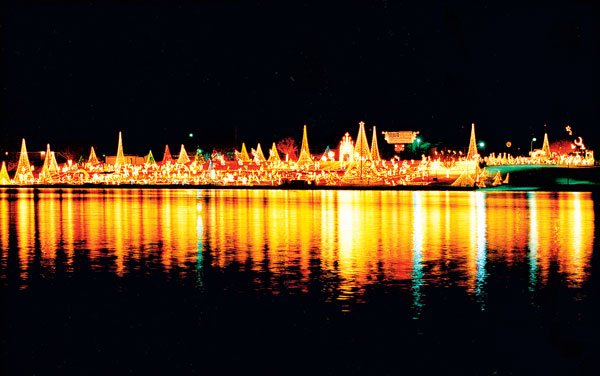 A vibrant nighttime scene showing illuminated boats decorated with bright lights reflecting on the water, creating a festive and colorful display against the dark sky.