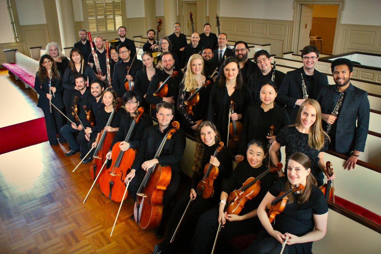 A group of musicians pose indoors with their string and wind instruments, smiling at the camera. They are arranged in rows within a spacious, well-lit room with wooden floors and cream-colored paneling.