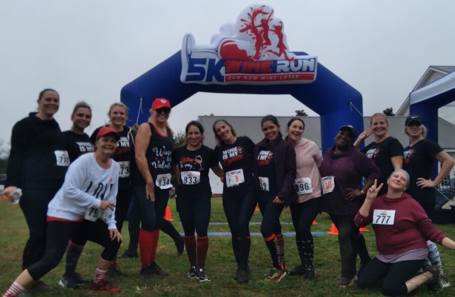A group of women poses and smiles in front of an inflatable arch that reads “5K Wine Run.” Most are wearing race bibs, athletic clothes, and fun socks, standing on grass with buildings and cones in the background.