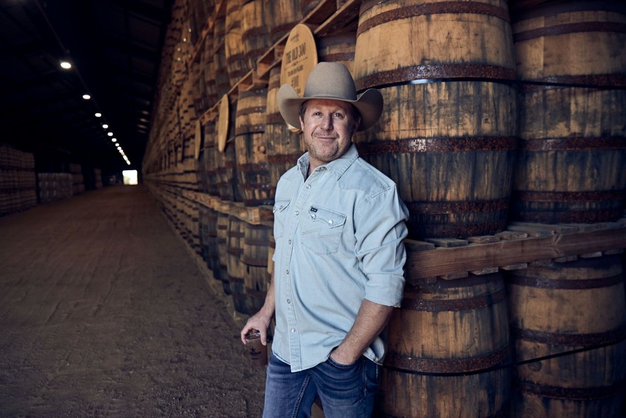 A man in a cowboy hat and denim shirt stands smiling with one hand in his pocket and the other holding a drink, leaning against stacked wooden barrels inside a large, dimly lit warehouse.