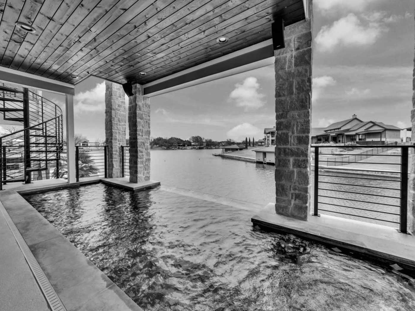 View from a covered patio of stunning vacation homes, featuring a small pool, stone pillars, and a wooden ceiling, overlooking a lake with houses and a dock in the background; spiral staircase on the left.