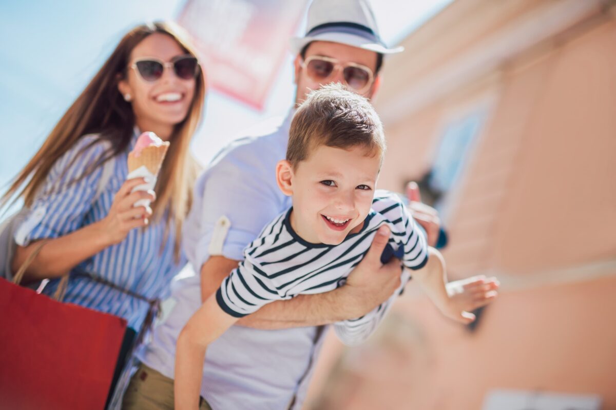 A smiling family enjoys a sunny day outside; an adult lifts a laughing young boy in the air while another adult smiles and holds an ice cream cone. All appear happy and relaxed.